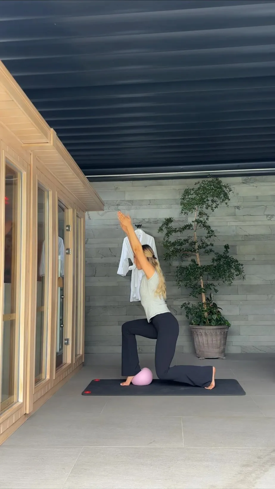 Charlotte on a mat outdoors, in a kneeling lunge with one arm raised, Pilates ball beneath her